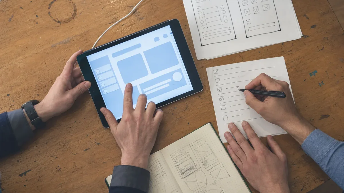 Two people reviewing a tablet mockup while checking off items on a written test checklist