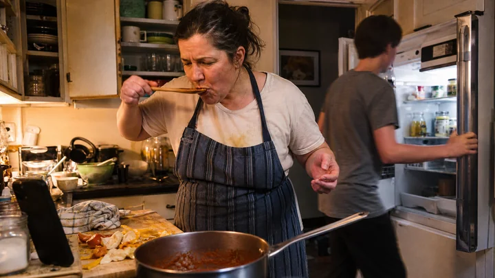 A cook tastes a family meal while another person checks ingredients in the refrigerator
